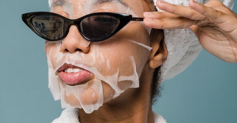 Cardio Routine - Self esteem young African American lady with moisturizing sheet mask and towel on hand wearing trendy sunglasses while standing against blue background in bathrobe after shower