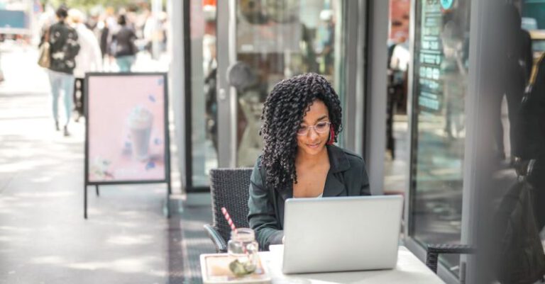 Online Programs - High angle of pensive African American female freelancer in glasses and casual clothes focusing on screen and interacting with netbook while sitting at table with glass of yummy drink on cafe terrace in sunny day